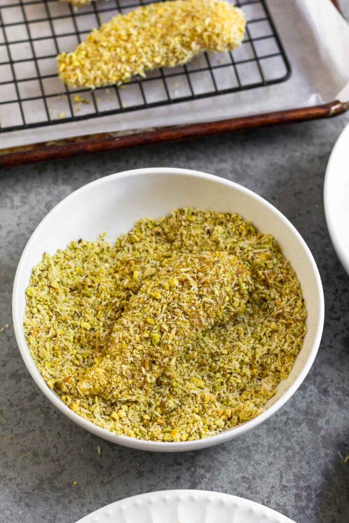 A bowl of breading with a chicken tender in it coated in it. Behind the bowl if a baking sheet with a wire cooling rack on it and a chicken tender that has already been breaded.