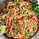 Plate filled with raw pad thai salad made from zucchini noodles, carrots, and bell peppers. It is garnished with lime wedges, cilantro, and peanuts. Next to it is a bowl of peanuts, white linen, and half a lime.
