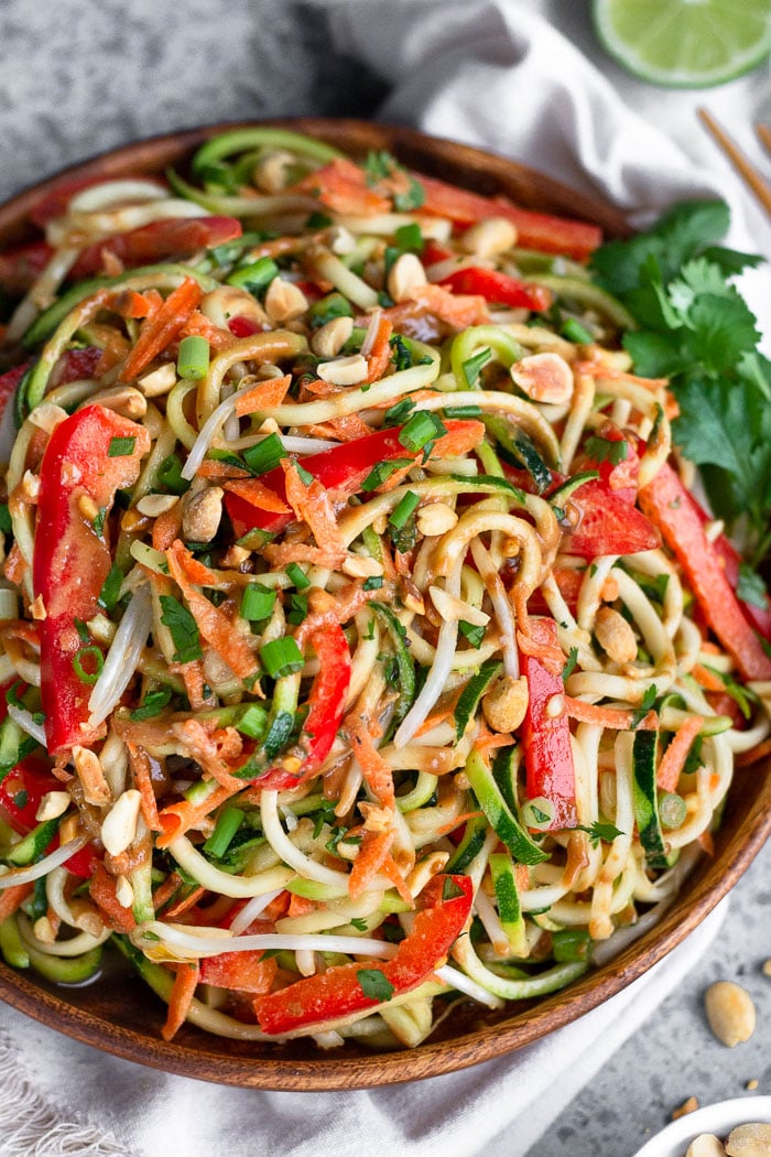 Wooden plate with vegan zucchini noodle pad thai garnished with cilantro, lime wedges, and chopped peanuts.