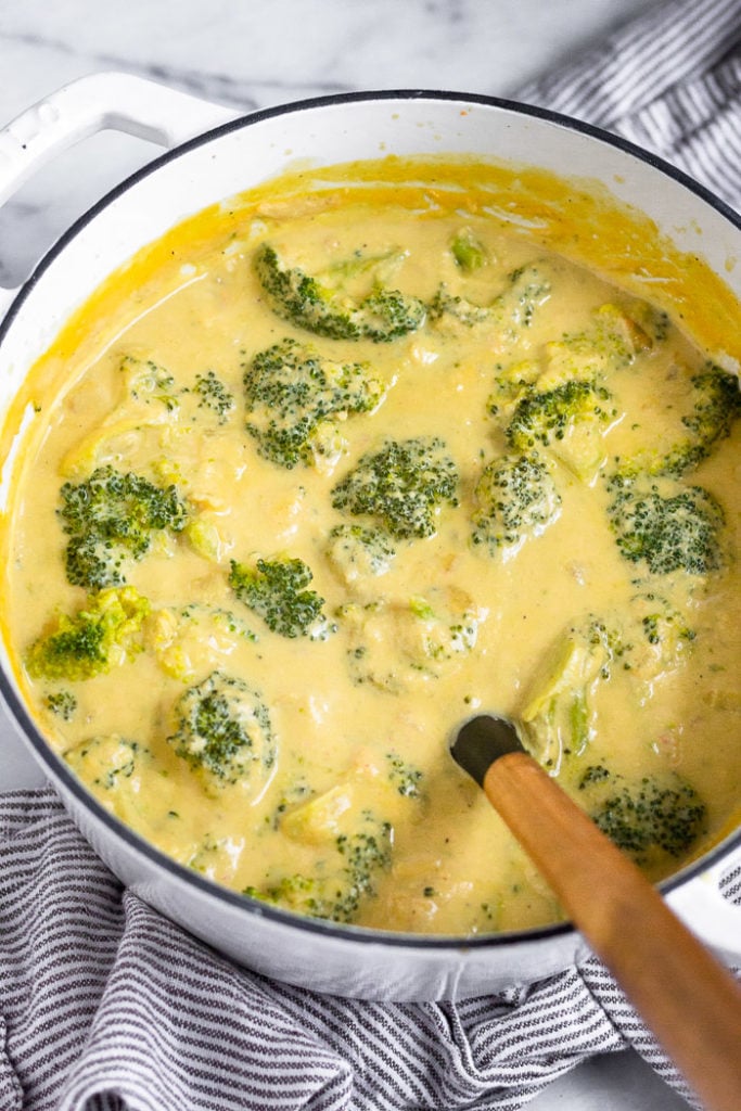 Vegan broccoli soup in a large white dutch oven with a wooden ladle sticking out of it. The pot is on a striped linen.