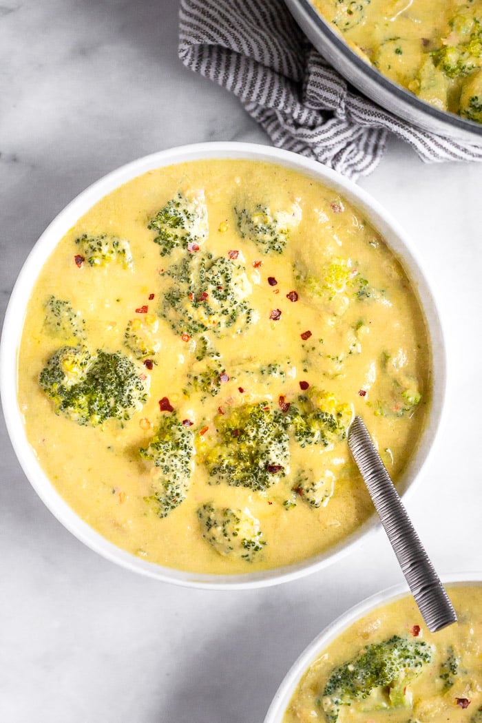 A bowl filled with vegan broccoli cheese soup topped with red pepper flakes and a spoon coming out of it. Next to it is another bowl of soup and a pot of soup.