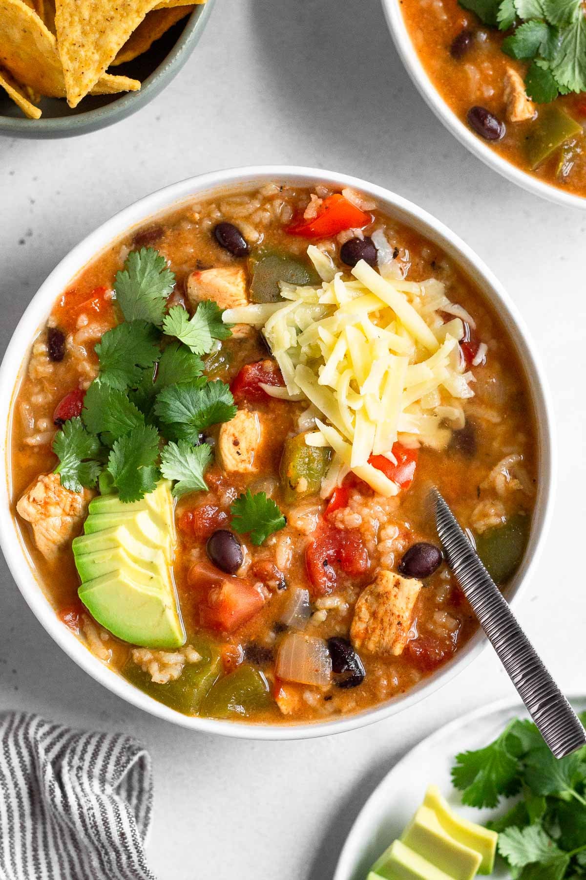 A bowl of chicken fajita soup topped with sliced avocado, cilantro, and shredded cheese. Around it is another bowl of soup, a bowl of tortilla chips, a striped kitchen towel, and a plate of sliced avocado and cilantro.