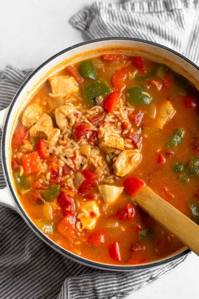 Large white dutch oven filled with chicken and rice soup with veggies. A wooden spoon is sticking out of it.