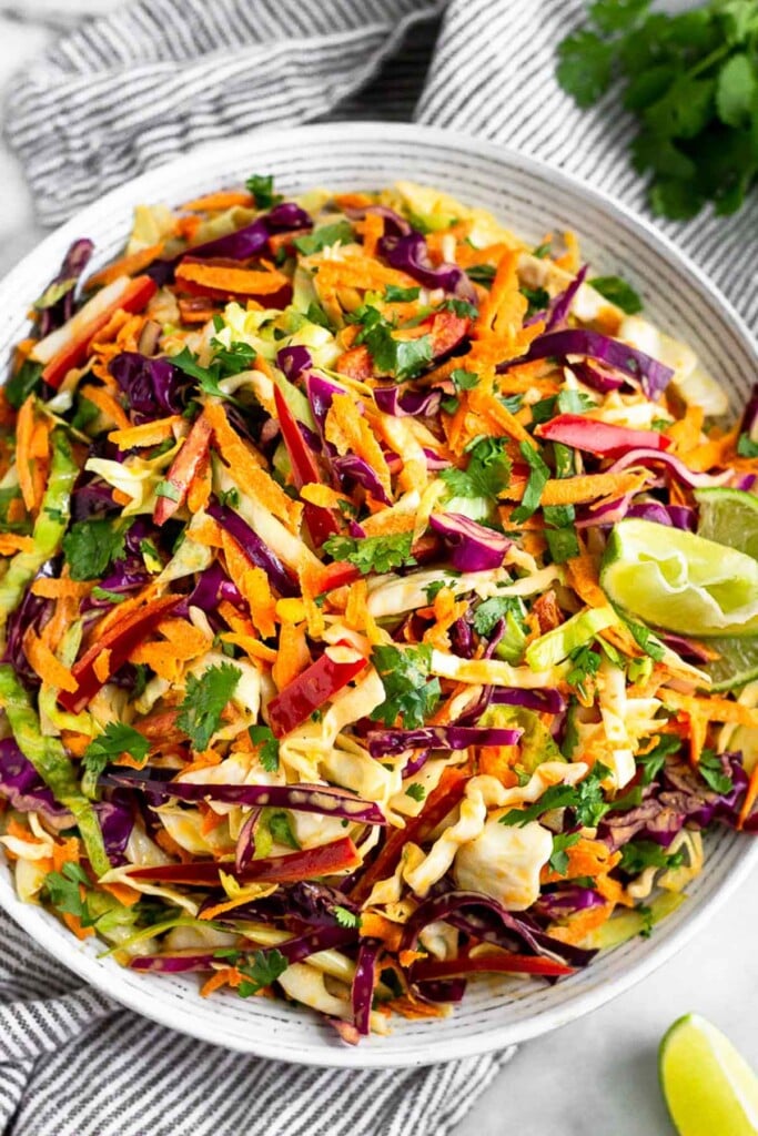 Large bowl of coleslaw with lime wedges on the side of the bowl. It is stilling on a striped towel with lime wedges and cilantro around it.