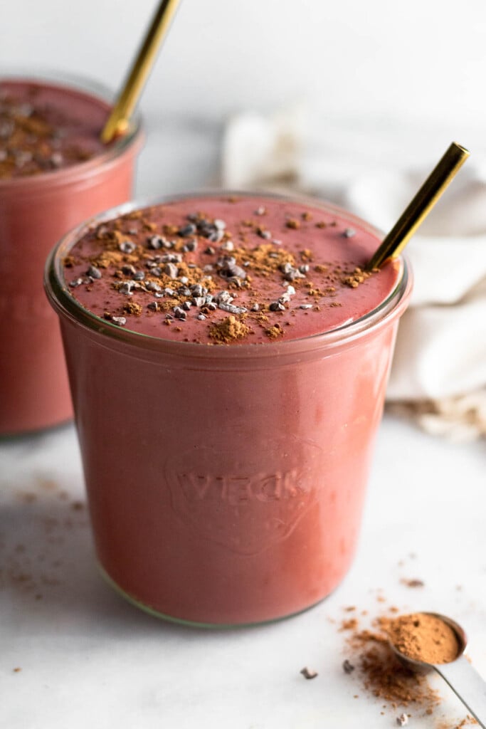 Two chocolate beet smoothies in jars topped with cacao nibs and gold straws in them. In front of them is a teaspoon filled with cacao powder and behind them is a tan linen.
