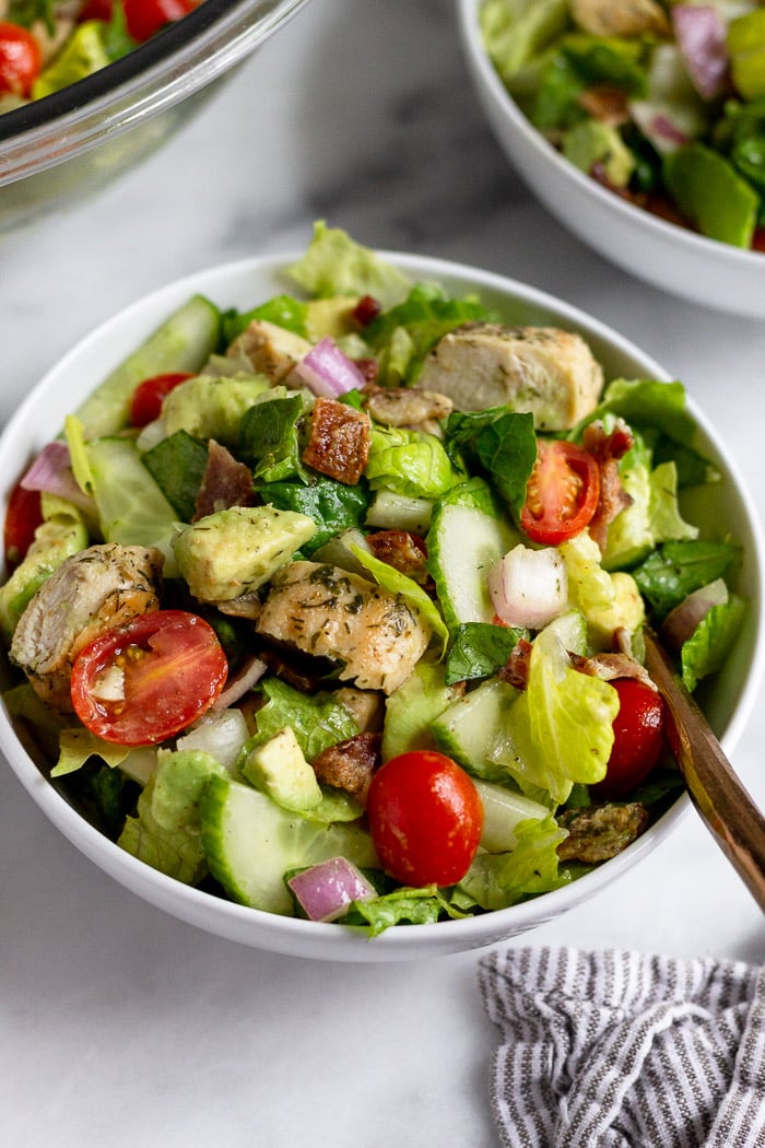 Bowl of chopped chicken salad with lettuce, tomatoes, cucumber, onion, and avocado with a fork sticking out of it,