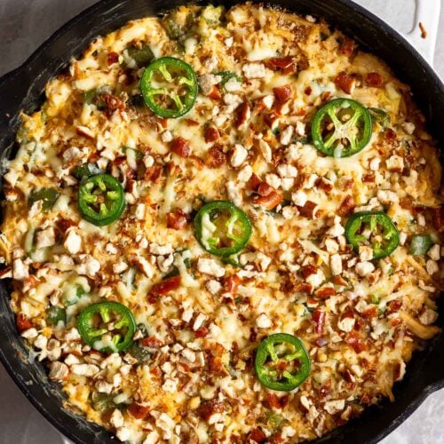 Cast iron skillet filled with jalapeño popper chicken casserole topped with melted cheese, crumbled pretzels, and sliced jalapeños. Next to it is a kitchen towel and bowl of chips.