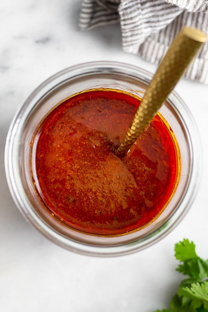 A jar of homemade red enchilada sauce with a spoon in it. Next to the jar is some cilantro and a kitchen towel.