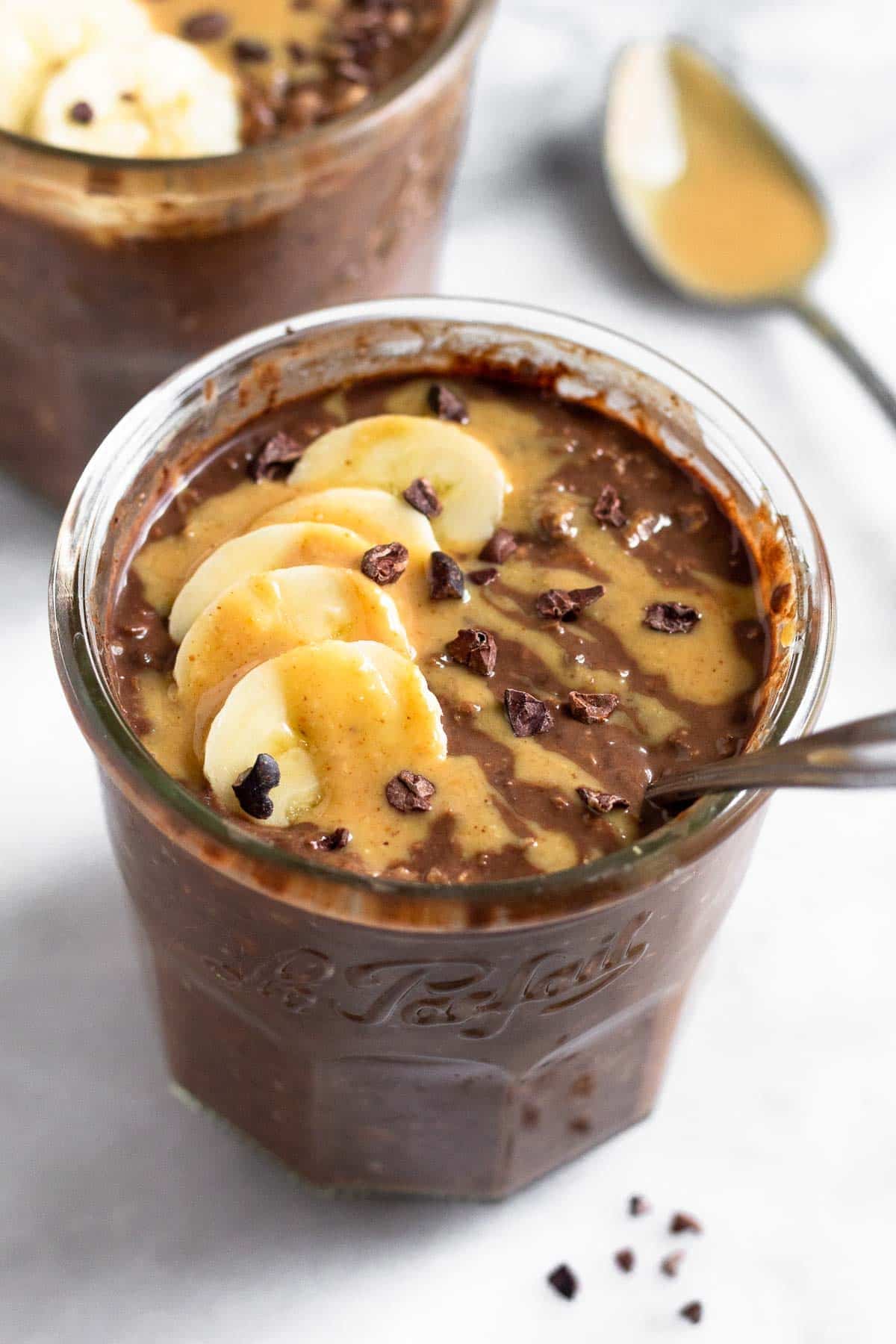 Chocolate peanut butter overnight oats in a jar topped with sliced banana, peanut butter, and cacao nibs with a spoon in the jar. Behind it is a another jar with overnight oats and a spoon with peanut butter on it.