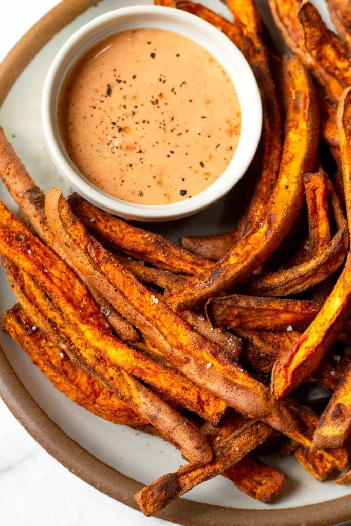 Close up of sweet potato fries sprinkled with salt on a white plate. On the plate is a small dish of dipping sauce.