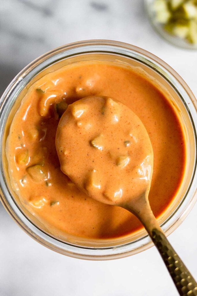 Burger sauce in a jar with a spoon overtop holding some. Off to the side is a bowl of diced pickles.