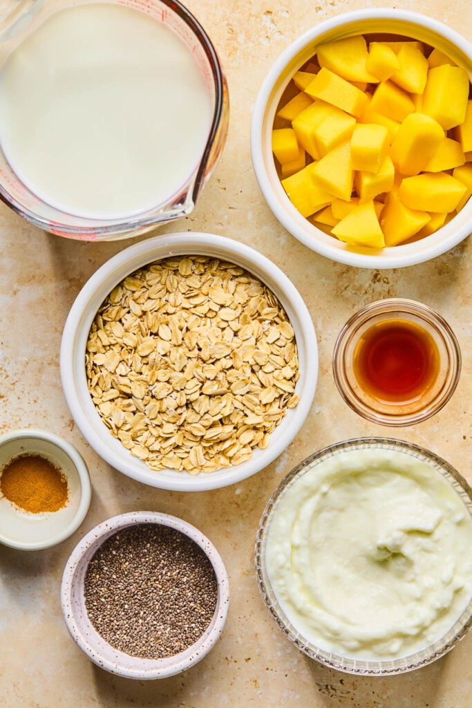 A bowl of diced mangos, a small bowl of vanilla extract, a bowl of greek yogurt, a bowl of chia seeds, a small bowl of cinnamon, and bowl of rolled oats, and a measuring cup of milk on a tan concrete counter.