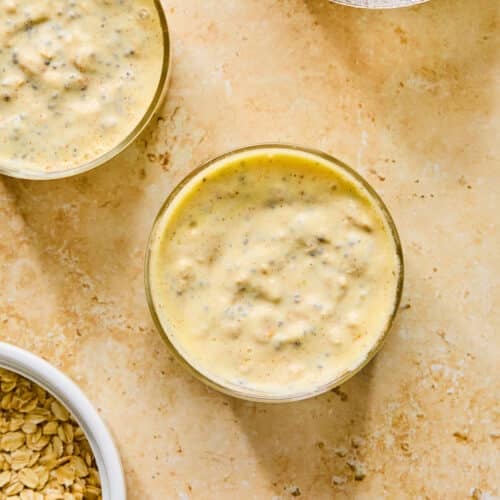 Overhead shot of blended mango overnight oats. Next to the jars are a small bowl of chia seeds, a bowl of rolled oats, and a bowl of greek yogurt.