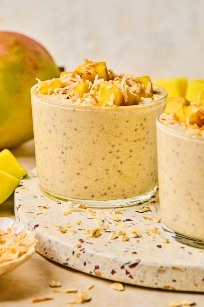 Two jars of overnight oats with mango on a terrazzo board. Next to the board is a small bowl of shredded coconut and a whole mango.