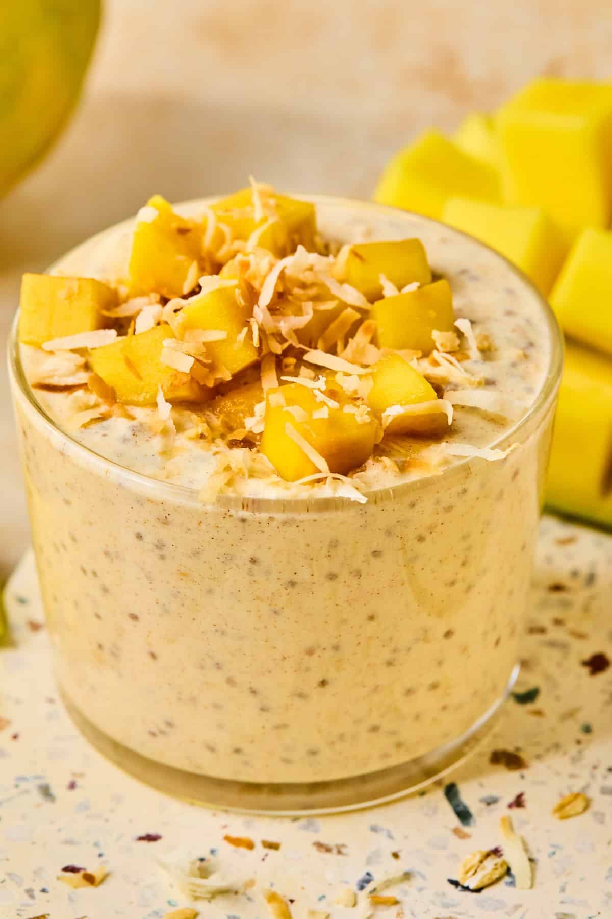 A jar of mango overnight oats topped with fresh diced mango and shredded coconut. Behind the jar is a cut mango.