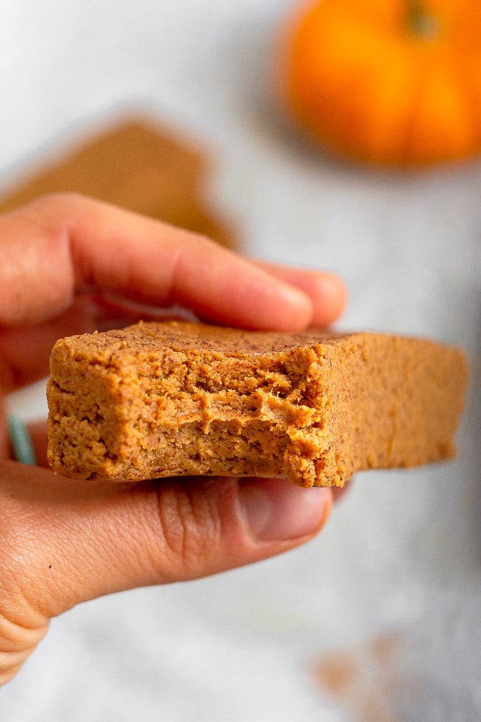 A hand holding a homemade pumpkin perfect bar with a bite taken out of it. In the background is a small pumpkin.