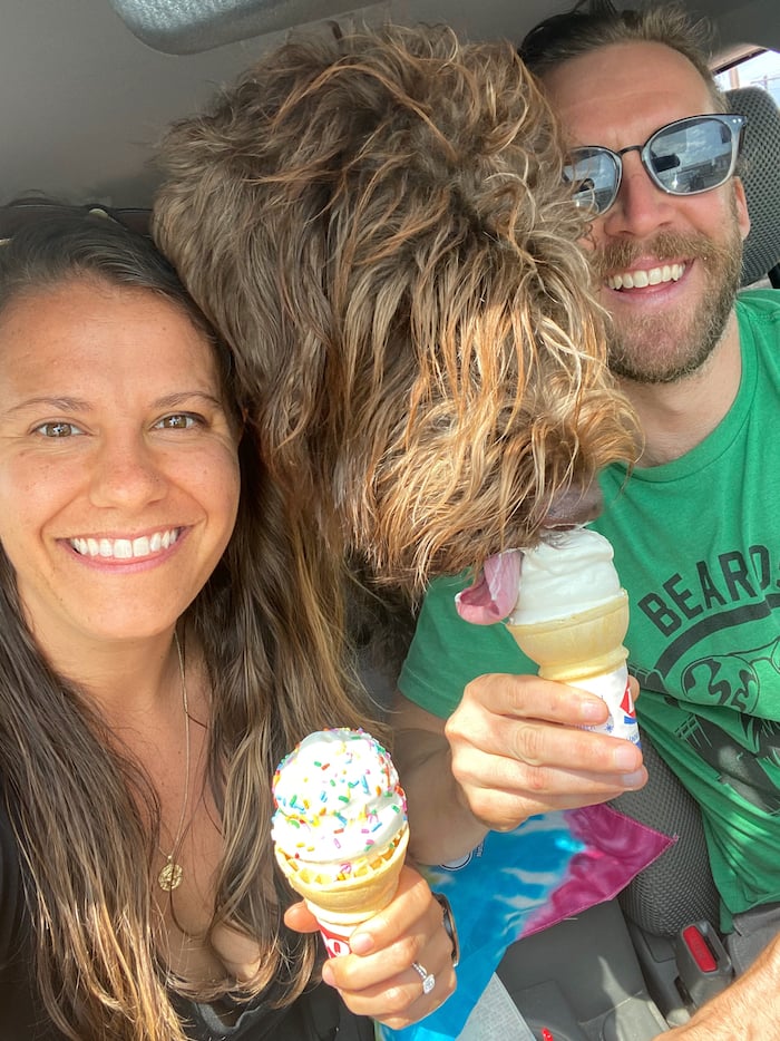 Woman and man both holding ice cream cones with a wirehaired pointing griffon dog between them licking the man's ice cream cone. 