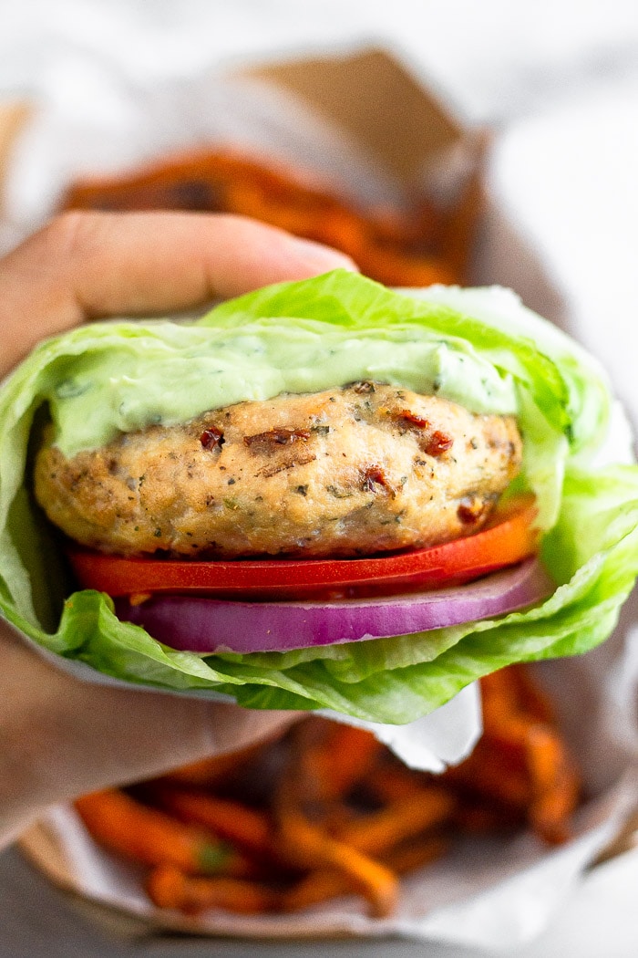 A hand holding a chicken burger with avocado aioli wrapped in a lettuce wrap. There is a pile of sweet potato fries behind it.