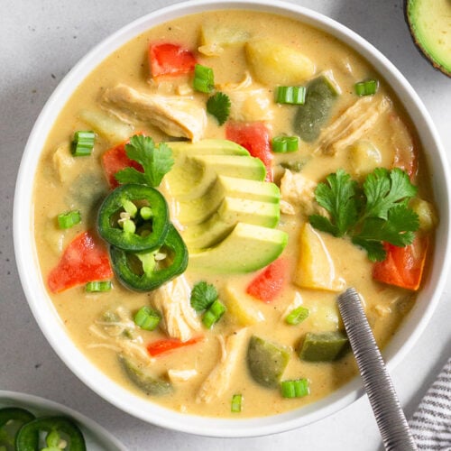 Overhead shot of cheesy chicken soup in a bowl topped with avocado, jalapeños, and herbs. A spoon is in the bowl and around it is a plate with more herbs, an avocado, a bowl of chips, and a kitchen towel.