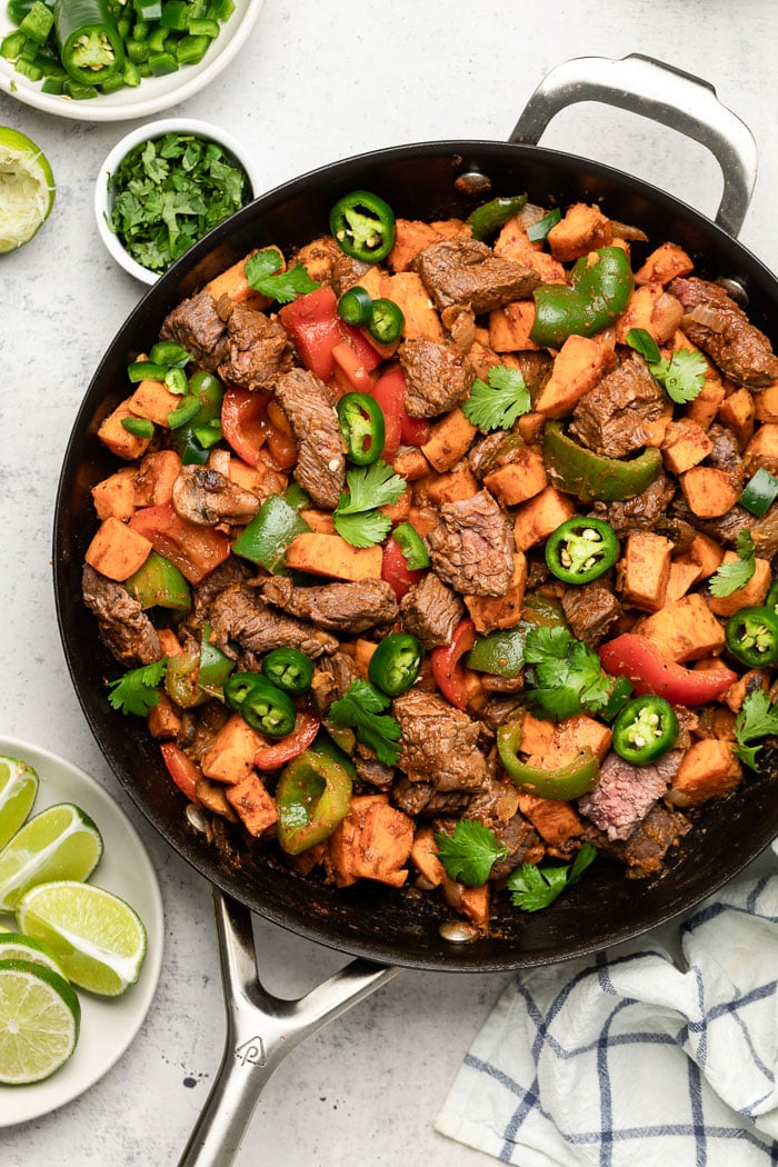 Steak and sweet potato skillet in a large pan topped with jalapeños and cilantro. Around it is a towel, lime wedges, and cilantro.