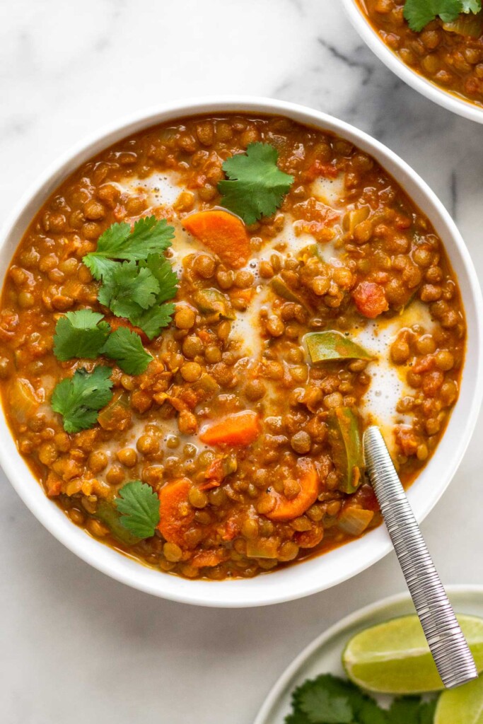 Curried lentil soup in a bowl topped with cilantro and a drizzle of coconut milk. A spoon is also in the bowl. Next to the bowl is another bowl of soup and a plate of lime wedges.