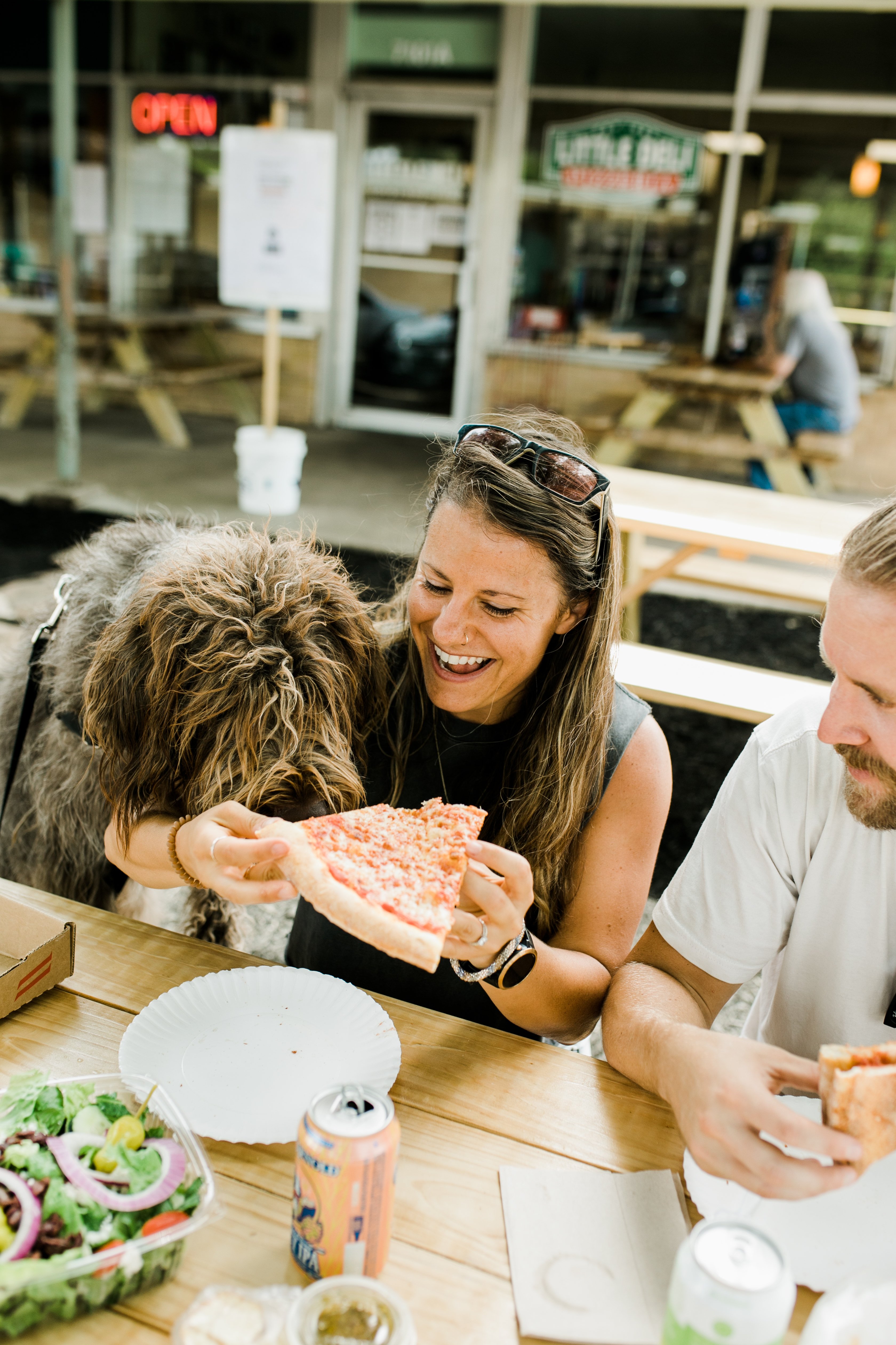 Female nutrition coach eating pizza with her dog and husband outside at a picnic table. She has a huge smile on her face.