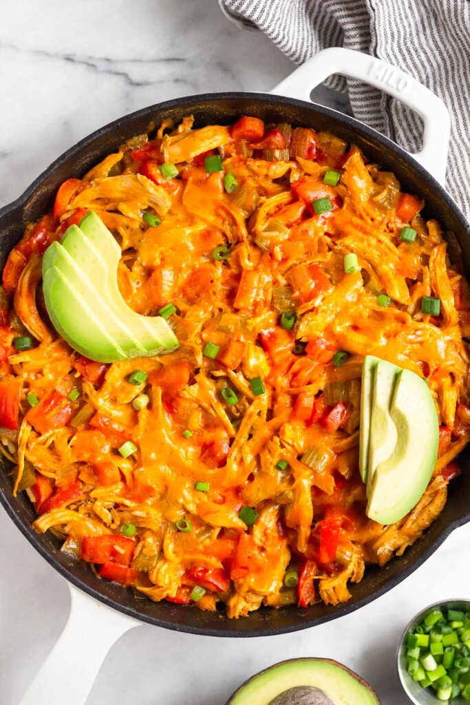 Buffalo chicken casserole topped with melted cheese in large cast iron skillet. It is garnished with sliced avocado and green onions. Next to it is a bowl of more green onions and half an avocado.