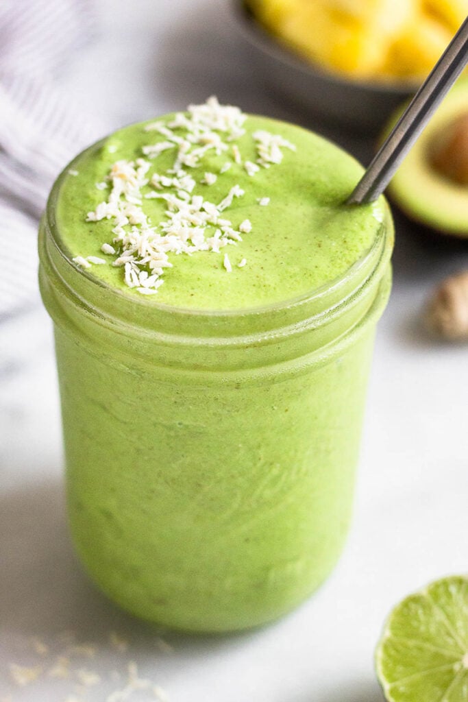 Green protein smoothie in a jar topped with coconut flakes and a metal straw in the jar. Around it is half a lime, a piece of ginger root, half an avocado, and a bowl of frozen pineapple.