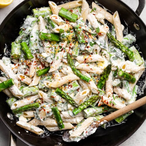 Large skillet filled with asparagus pasta in a creamy feta sauce. It is topped with lemon zest and red peppers flakes and a wooden spoon is in the bowl. Around the pan is a dish of chopped herbs, some lemons, a bowl of feta, and a small dish of red pepper flakes.