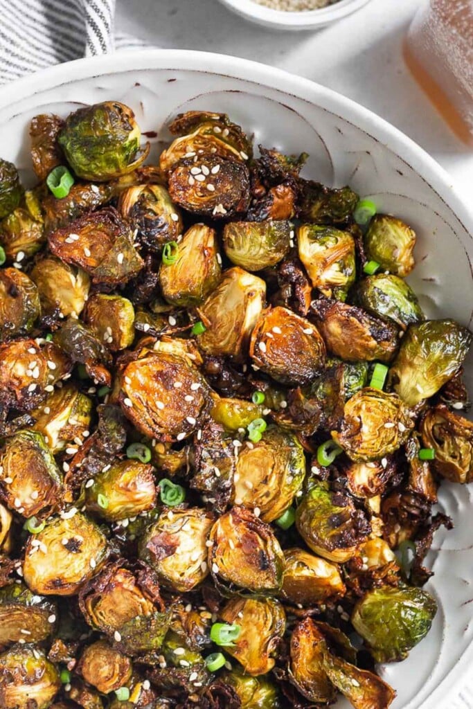 Crispy roasted brussel sprouts garnished with sesame seeds and green onions in a white bowl. Next to the bowl is a jar of honey and a striped towel.