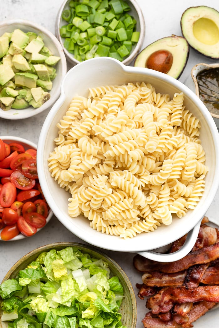 Overhead shot of a bowl of cooked pasta, plate of cooked bacon, bowl of chopped lettuce, bowl of cherry tomatoes, bowl of avocado, bowl of green onion, and some halved avocado and herbs.