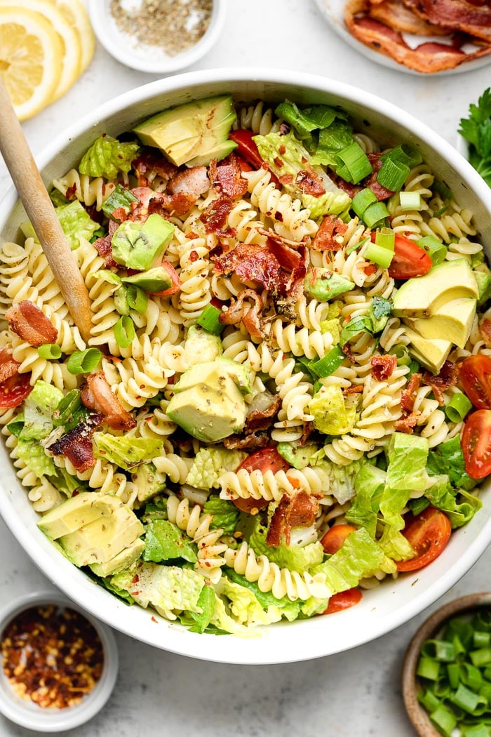 BLT pasta salad with Italian dressing and bacon in a large white bowl with a wooden spoon coming out of it. Around it is a few dishes of herbs, lemon, spices, and green onions.