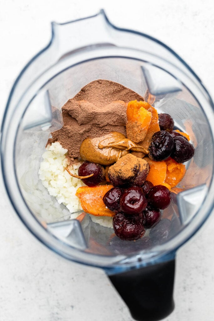 Overhead shot of a blender with frozen cauliflower rice, frozen sweet potatoes pieces, frozen cherries, chocolate protein powder, almond butter, and cinnamon in it.