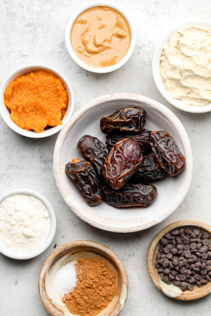 Grey counter with a bowl of nut butter, a bowl of vanilla protein powder, a bowl of medjool dates, a bowl of mini chocolate chips, a bowl of spices, a bowl of flour, and a bowl of pumpkin puree.