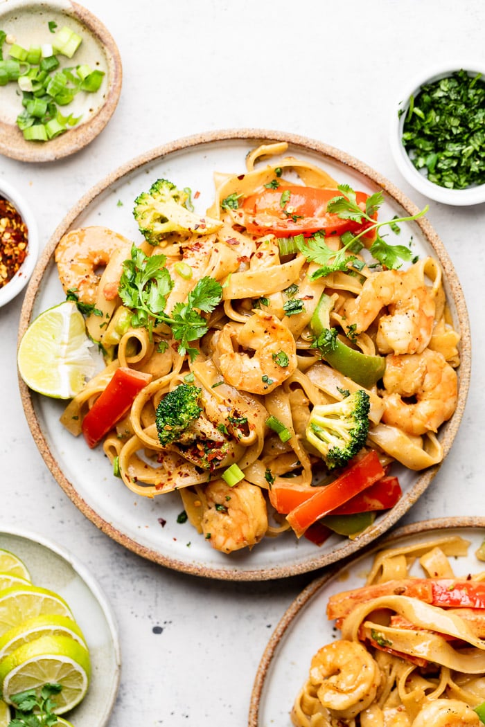 Plate of shrimp rice noodles with veggies and peanut sauce. It is topped with cilantro and has a lime wedge on the plate. Around it is another plate with shrimp noodles, small plate with lime wedges, bowl of red pepper flakes, bowl of chopped green onion, and a bowl of cilantro.
