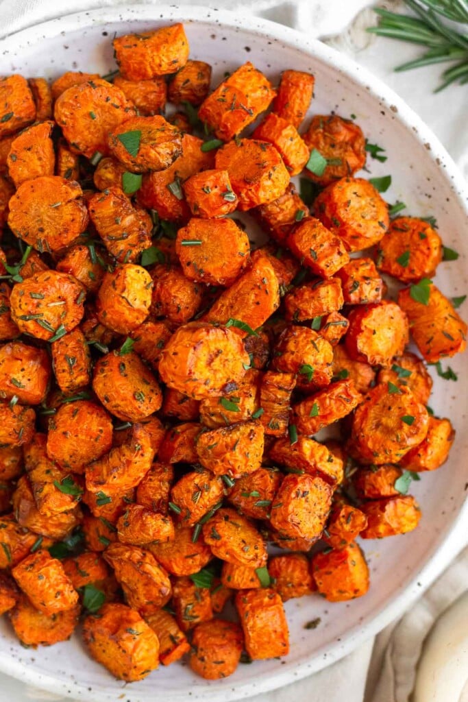 Air fryer roasted carrots on a white plate topped with fresh herbs. There is some fresh rosemary next to the plate.