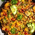 Overhead shot of chicken fajita casserole topped with melted cheese in large cast iron skillet. It is garnished with sliced avocado, sliced jalapeños, and cilantro. Around the pan is striped kitchen towel, a bun of cilantro, and lime wedges.