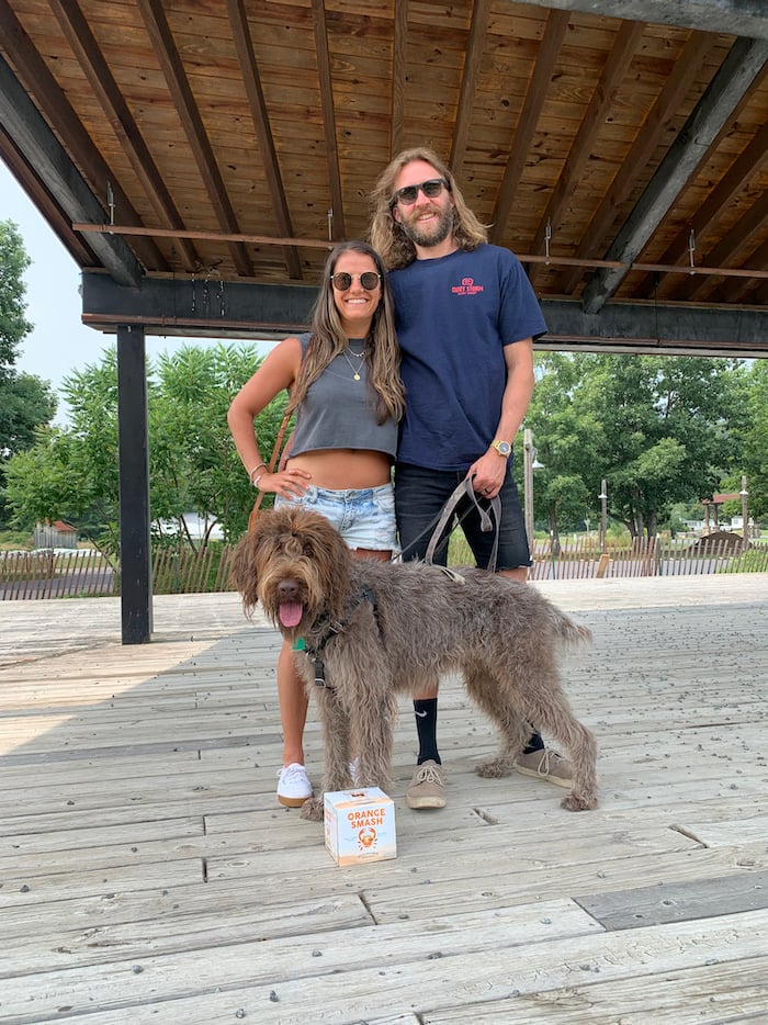 Husband and wife and their wirehaired pointing griffon dog on a stage with trees in the background.
