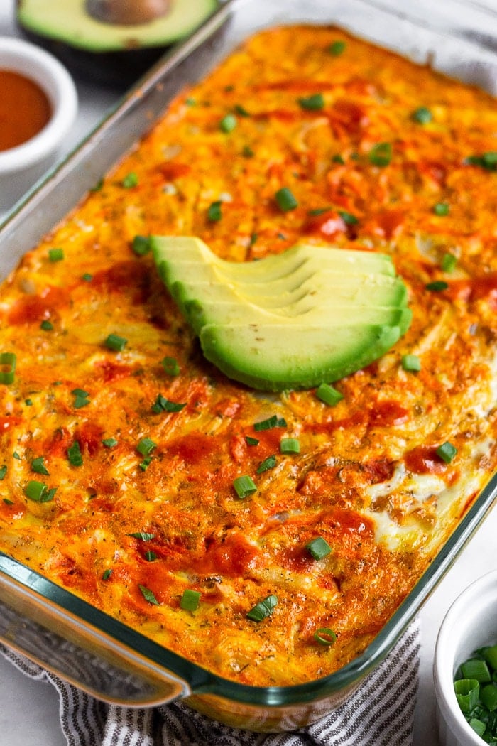 Chicken breakfast casserole in a casserole dish topped with avocado, hot sauce, and green onions. Next to it a ramekin of chopped green onions, ramekin of buffalo sauce, and half an avocado.