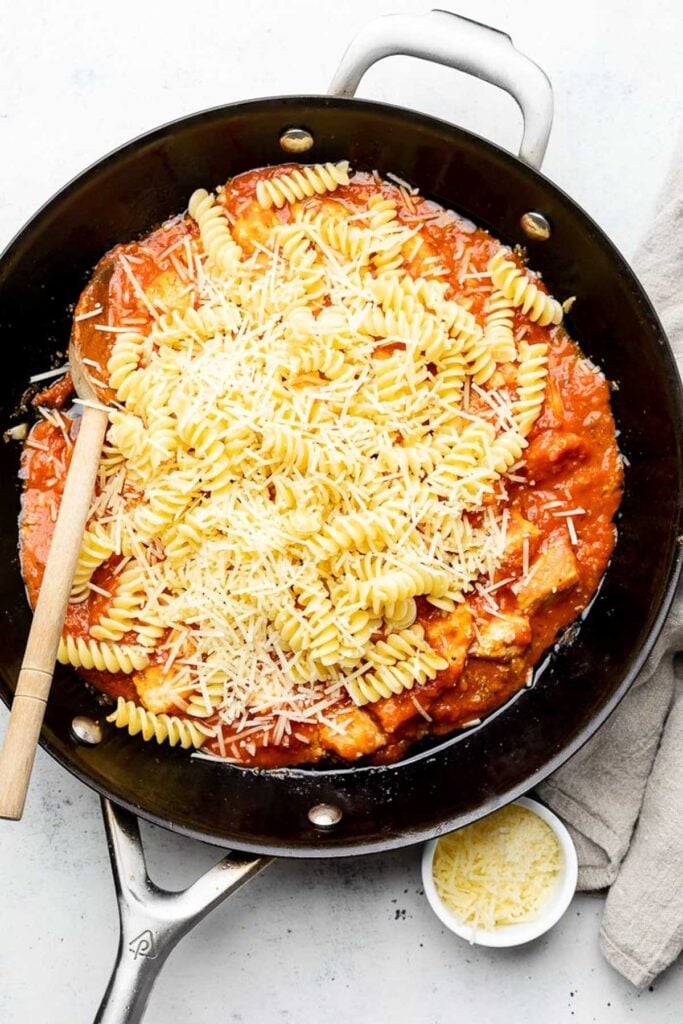 Skillet filled with cooked diced chicken mixed with marinara sauce. On top of that is a pile of cooked pasta and shredded parmesan cheese.