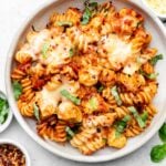 Chicken parmesan pasta in a bowl topped with fresh basil. Next to the bowl is more fresh basil, a bowl of red pepper flakes, and a bowl of parmesan cheese.