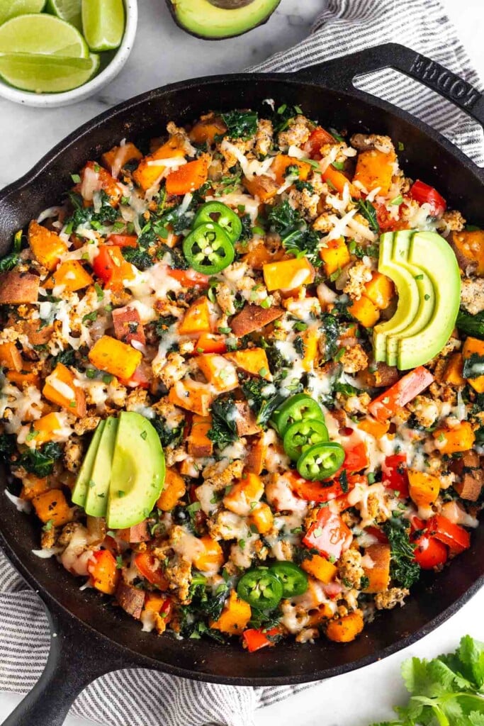Ground turkey skillet with sweet potatoes, peppers, and kale in a large cast iron skillet. It is garnished with sliced avocado, sliced jalapeno, and cilantro. Around the skillet is a bowl of lime wedges, half an avocado, and fresh cilantro.