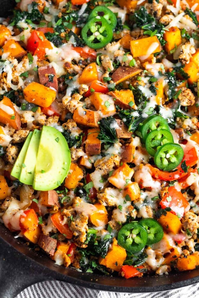Ground turkey, sweet potatoes, and veggies in a large skillet topped with melted cheese. It is garnished with sliced avocado and sliced jalapeños.