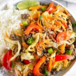 A plate of ground beef and cabbage stir fry with white rice and a lime wedge. It is garnished with sesame seeds and green onions. Behind the plate is another plate and a bowl of sesame seeds.
