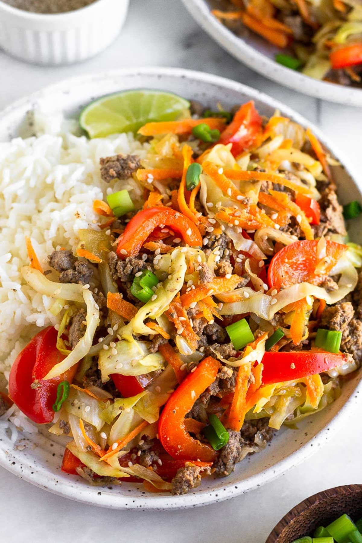 A plate of ground beef and cabbage stir fry with white rice and a lime wedge. It is garnished with sesame seeds and green onions. Behind the plate is another plate and a bowl of sesame seeds.