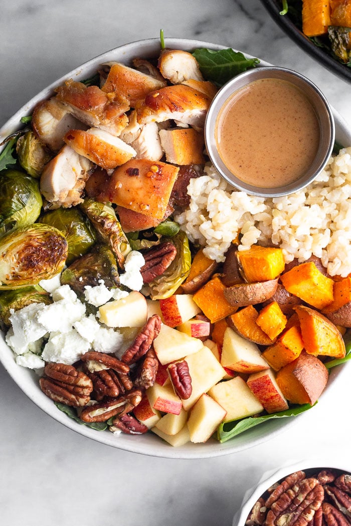 Healthy bowl recipe with chicken, rice, sweet potatoes, apples, nuts, cheese, and brussel sprouts with a small ramekin of dressing in a bowl.