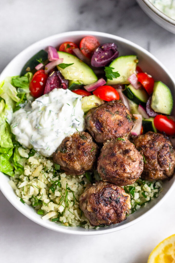 Healthy bowl recipe with cauliflower rice, greek salad, lamb meatballs, and tzatziki dressing in a bowl.