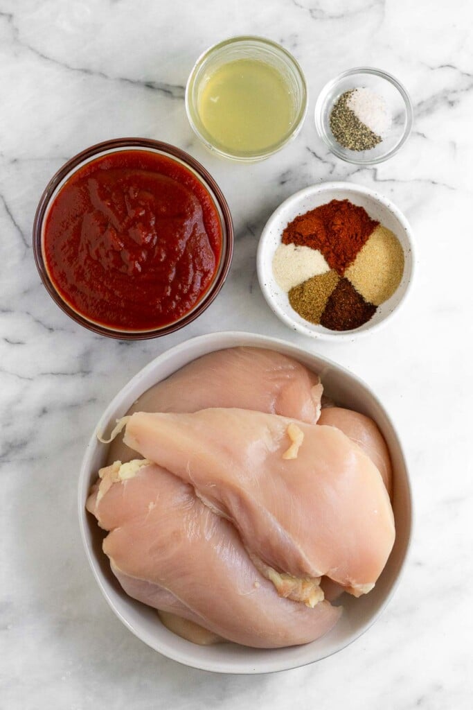 White marble counter with a jar of chicken broth, a small bowl of salt and pepper, a bowl of different spices, a large bowl of raw chicken breasts, and a bowl of barbecue sauce.