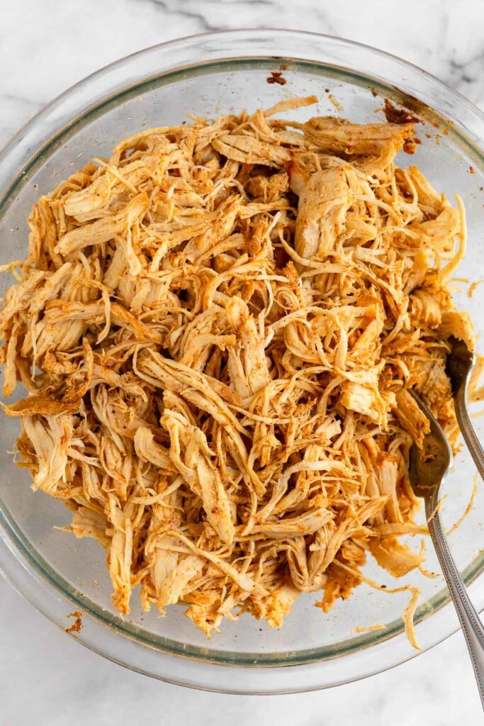 A glass bowl of seasoned shredded chicken with two forks in it.