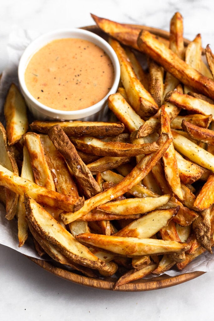 Homemade air fryer french fries on a plate sprinkled with salt. There is also a small container of orange sauce on the plate.